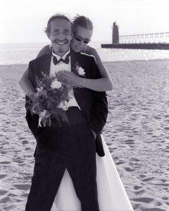 Penn street hugging her husband from behind on a beach on their wedding day.
