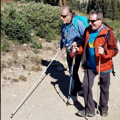 A volunteer guiding a man with a white cane on a hiking trail.
