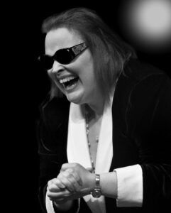 Black and white photo of diane standing, laughing, with hands together. Wearing glasses and suitejacket.