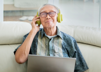 Senior man with a laptop sitting on a couch, with headphones on, smiling