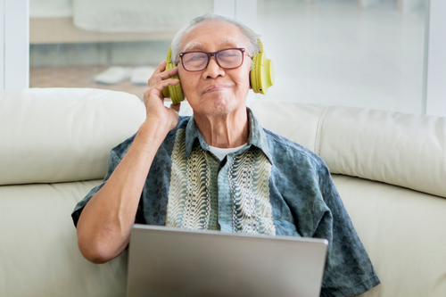 Senior man with a laptop sitting on a couch, with headphones on, smiling