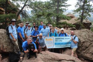 Group of several people standing in boulders on a mountain, smiling and holding an AINC banner.