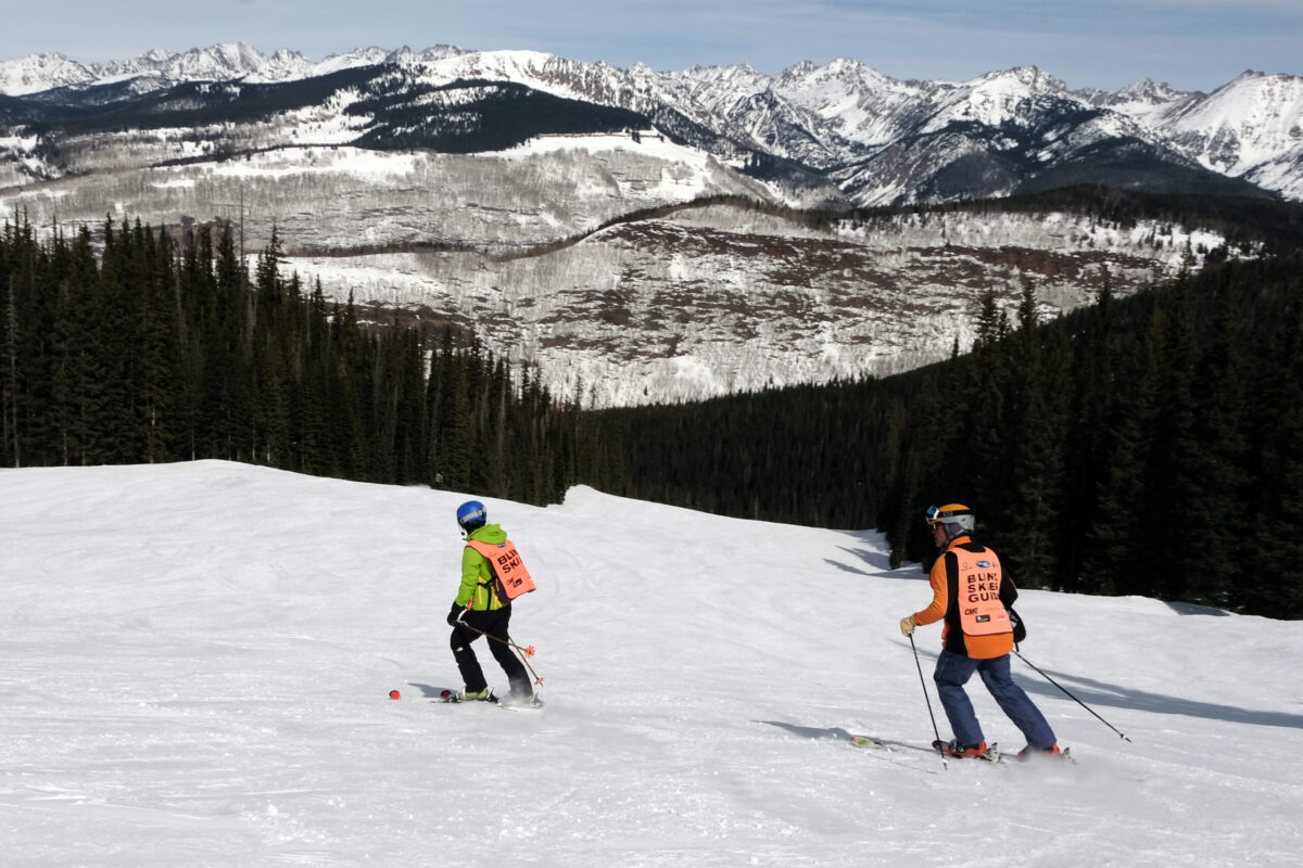Two skiers wearing orange vests on a slope with mountain range in the background