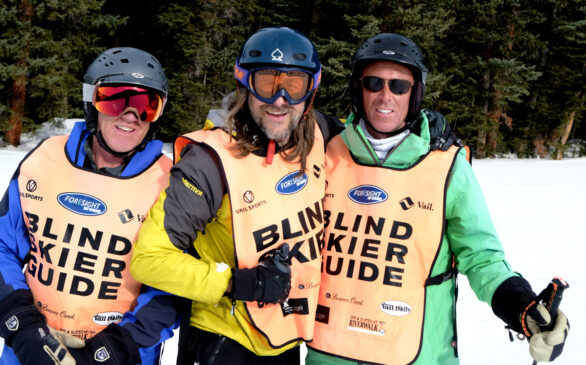 Three skiers laughing together, with orange vests that say "Blind skier" and "Blind Skier guide"